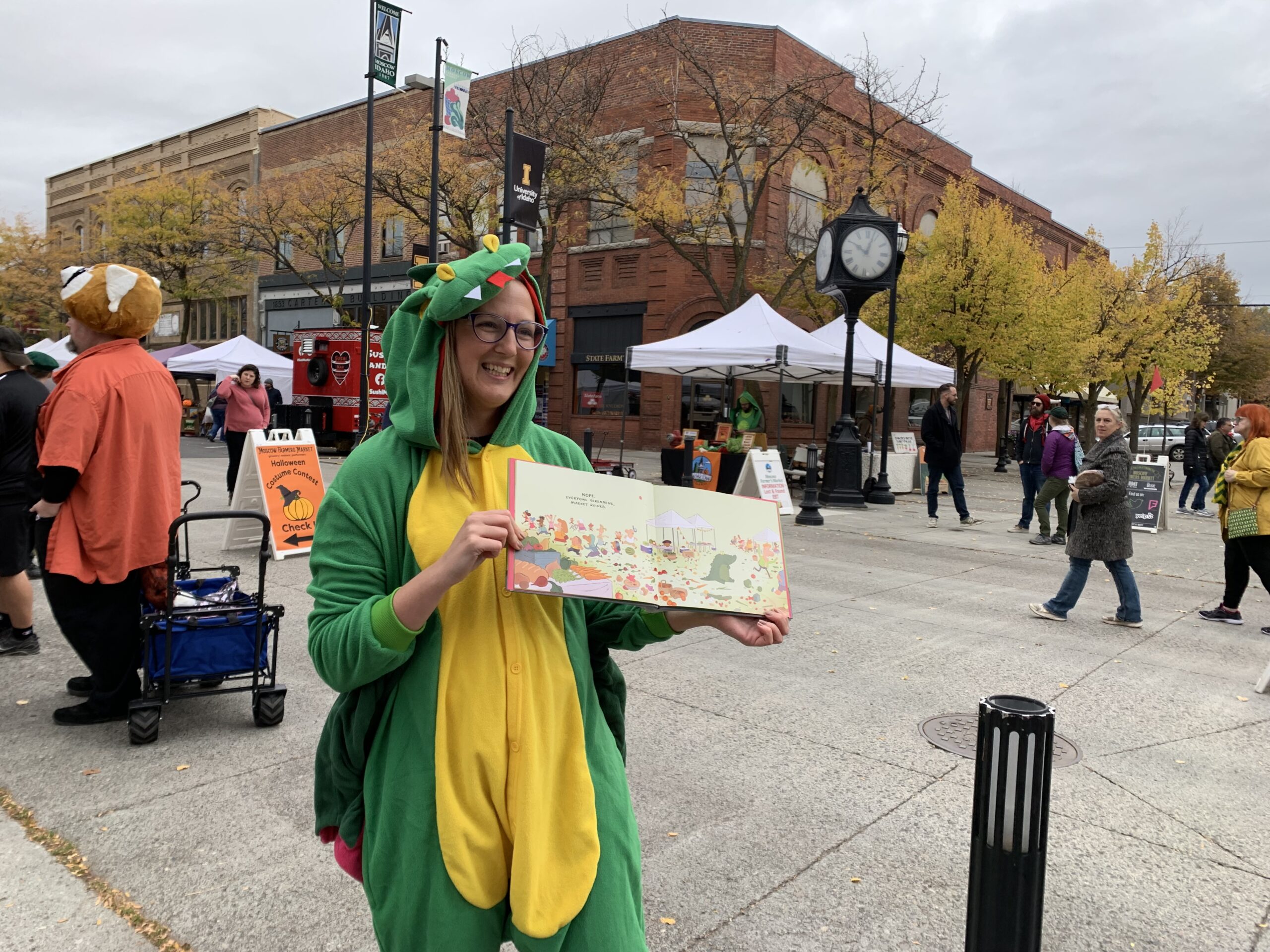 Eija wearing a crocodile costume while holding her book open to the farmer's market page during an October farmer's market overcast morning.