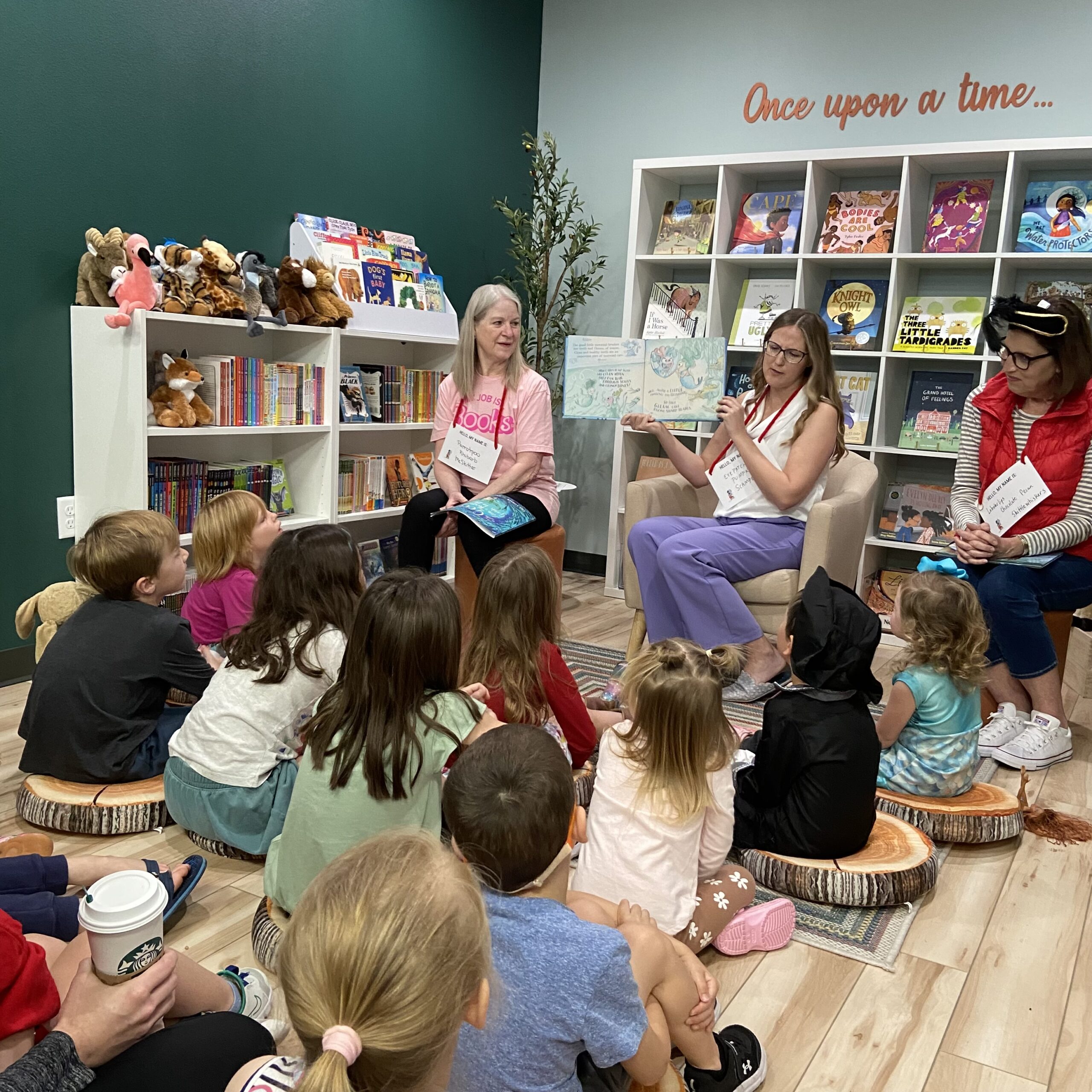 Storytime in a bookshop with dark green and light green walls, filled with bookshelves, children, and storytime friends.